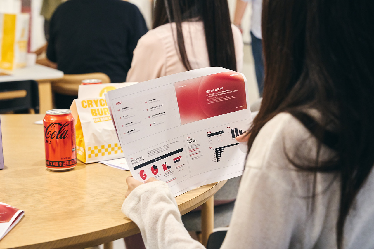 차트가 실린 인쇄물을 펼쳐 든 참석자와 테이블 위 콜라 캔, 버거 봉투